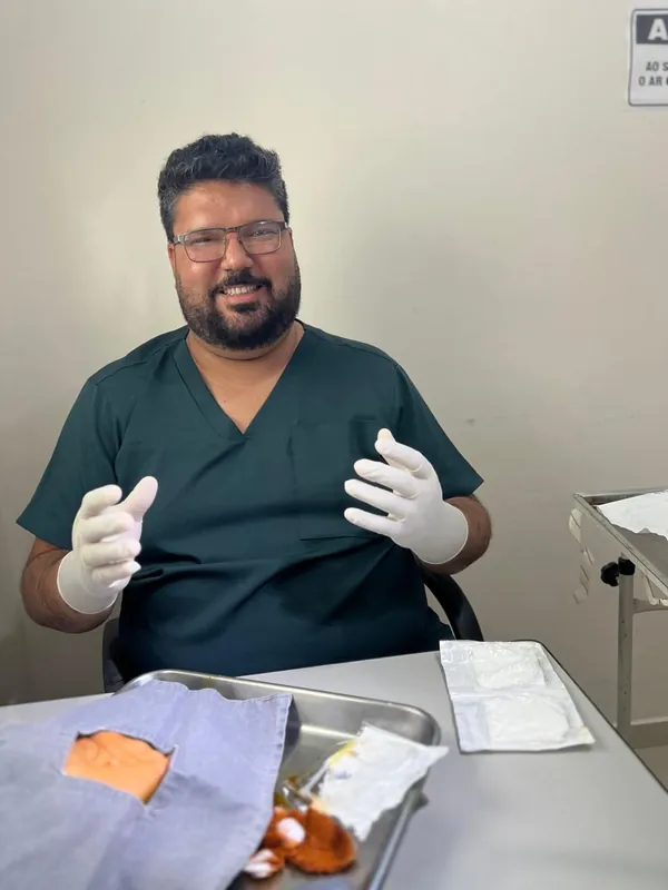 Dr. Sérgio de luvas cirúrgicas, sorrindo, ao lado da bandeja de um procedimento